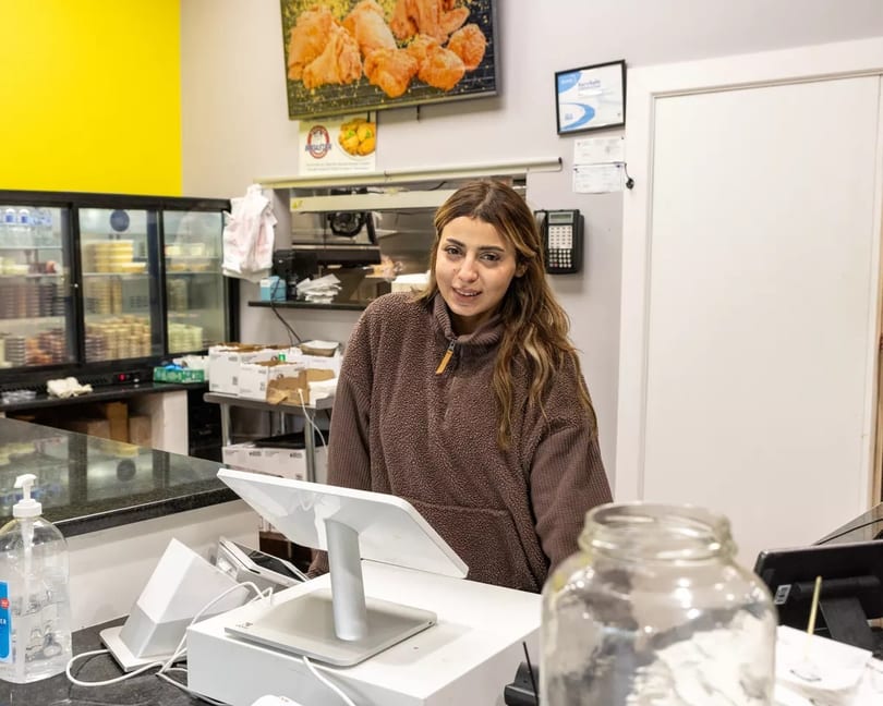 A smiling woman in a brown fleece stands behind a counter with a POS system.