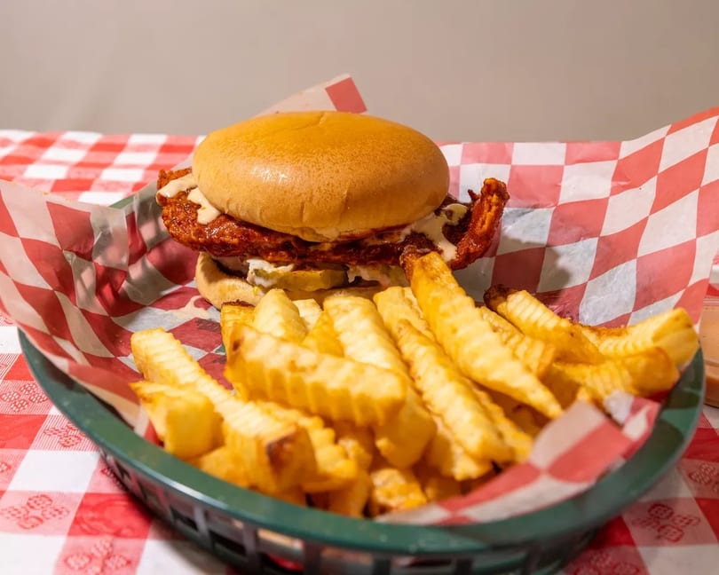 Spicy fried chicken sandwich with pickles and sauce, served with crinkle-cut fries in a basket.
