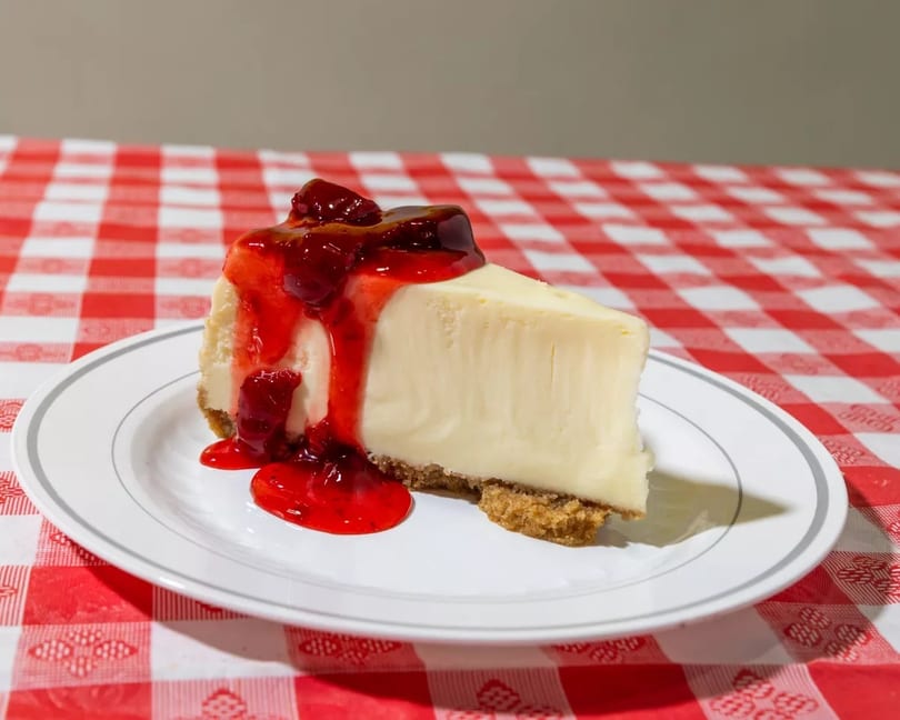 Slice of cheesecake with strawberry topping on a white plate, on a red and white checkered tablecloth.